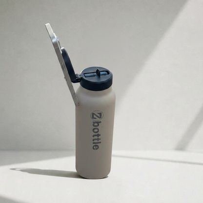 Botella beige con tapón negro, soporte para teléfono y grabado metálico sobre fondo fotográfico blanco