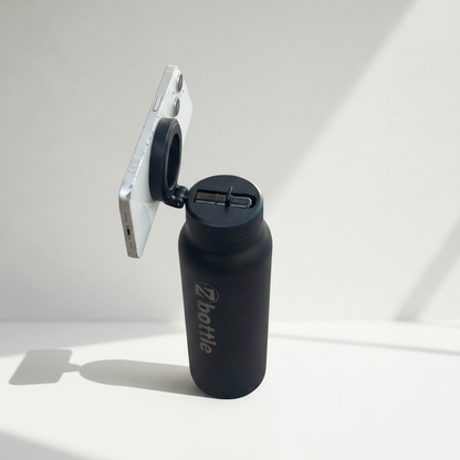 Botella negra con tapón negro, soporte para teléfono y grabado metálico sobre fondo fotográfico blanco